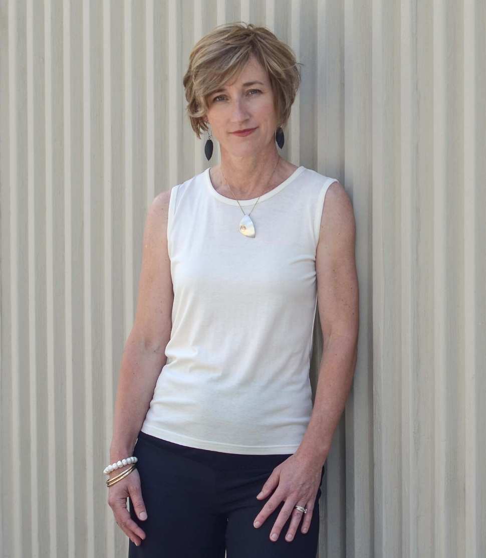 Woman wearing a Natural White Sleeveless Crew and black pants against a corrugated metal background