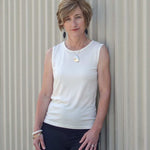 Woman wearing a Natural White Sleeveless Crew and black pants against a corrugated metal background