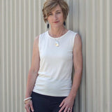 Woman wearing a Natural White Sleeveless Crew and black pants against a corrugated metal background